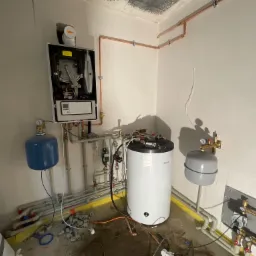 Montaż kotła gazowego Vaillant z podłączeniem do instalacji grzewczej i wodnej, widoczne rury miedziane, zbiornik wyrównawczy i manometry w surowym pomieszczeniu.