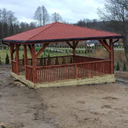 Nowo wybudowana drewniana altana z czerwoną dachówką bitumiczną, częściowo ogrodzona drewnianymi barierkami, na tle ogrodu i pochmurnego nieba.