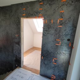 Mom-Bud Usługi Remontowo-Budowlane Łukasz Mortel - Ściana w trakcie układania płytek o ciemnej, wzorzystej fakturze, z pomarańczowymi krzyżykami dystansowymi i widocznymi opakowaniami płytek pod ścianą, w tle jasne pomieszczenie z oknem dachowym.