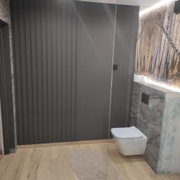Mom-Bud Usługi Remontowo-Budowlane Łukasz Mortel - Nowoczesna łazienka z szarą ścianą z pionowymi panelami, podwieszaną toaletą, marmurowym wykończeniem i drewnopodobną podłogą, uzupełniona okrągłym dywanikiem i szklaną ścianką prysznicową.
