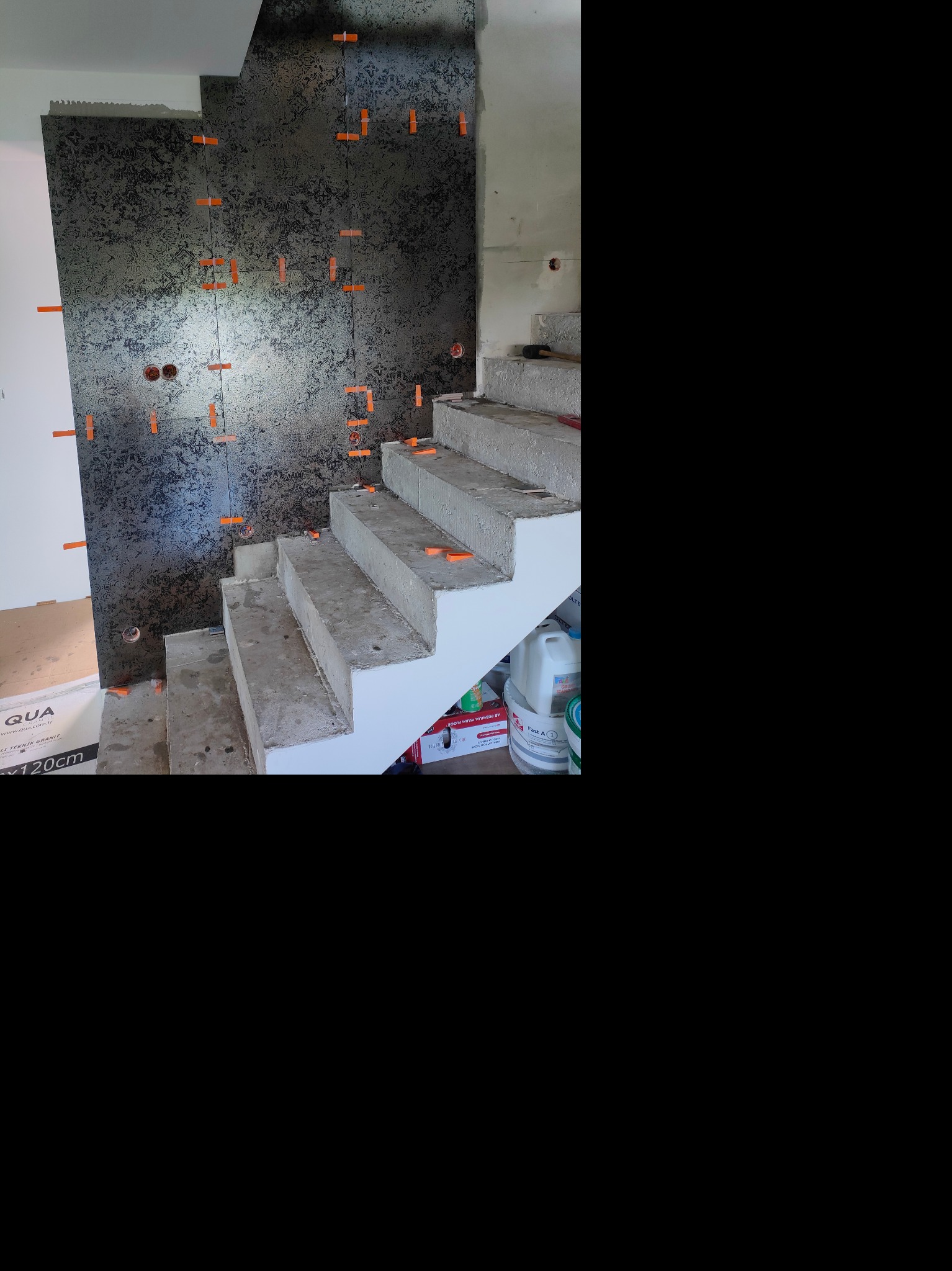 Montaż dekoracyjnych płytek ściennych w odcieniach szarości z użyciem pomarańczowych krzyżyków dystansowych, widoczne surowe schody betonowe w trakcie wykończenia.