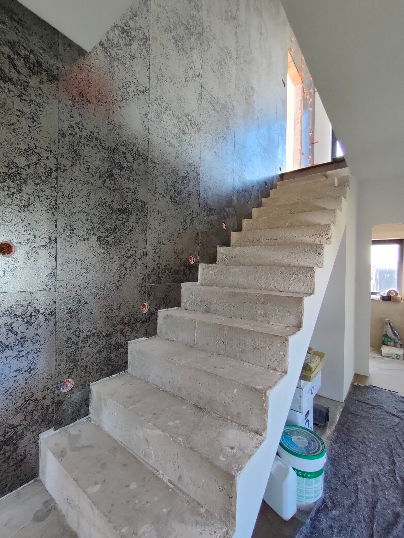 Surowe betonowe schody wewnętrzne z dekoracyjną ścianą w odcieniach szarości, widoczne puszki instalacyjne, w tle okno i materiały budowlane.