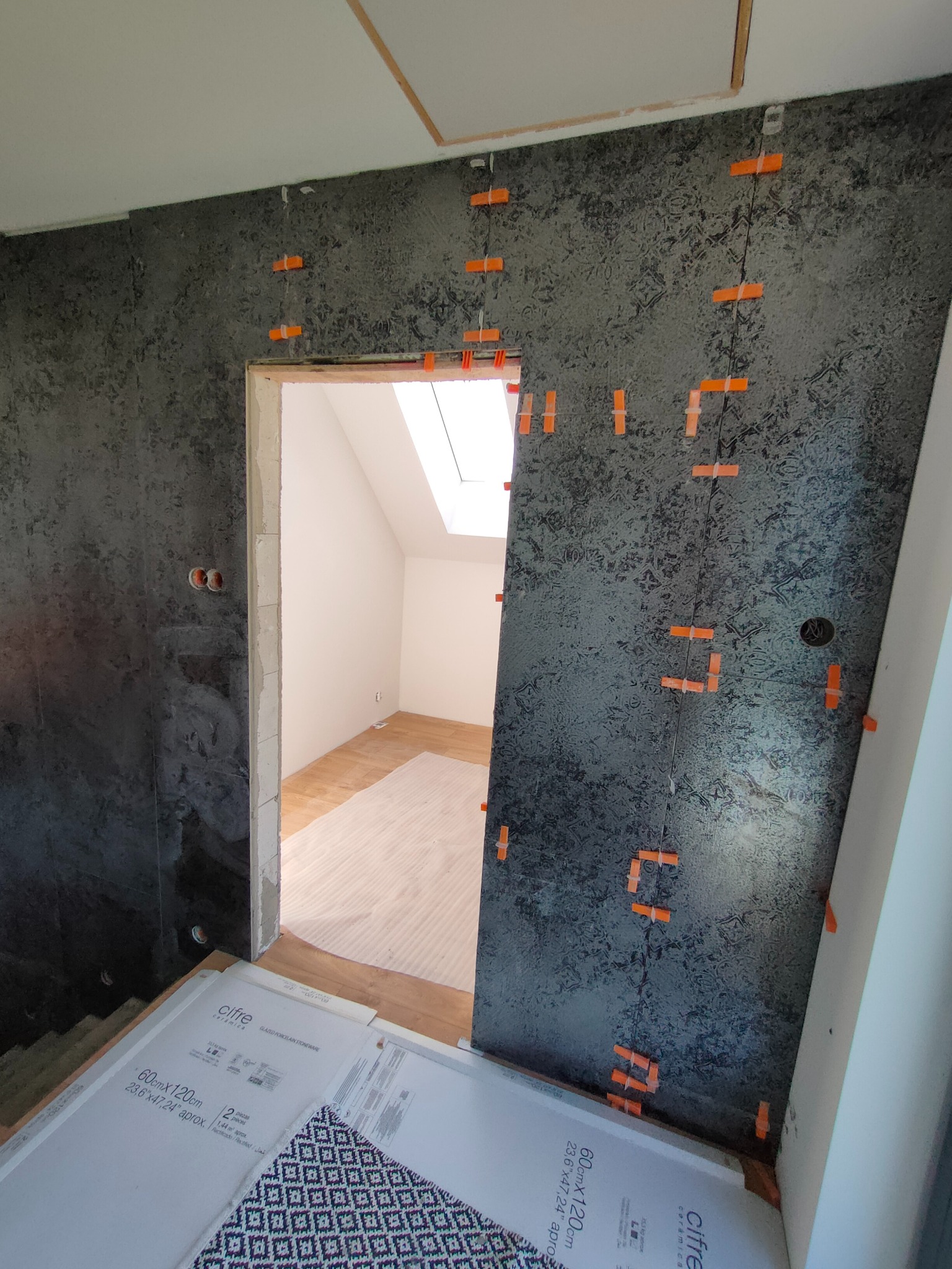 Ściana w trakcie układania płytek o ciemnej, wzorzystej fakturze, z pomarańczowymi krzyżykami dystansowymi i widocznymi opakowaniami płytek pod ścianą, w tle jasne pomieszczenie z oknem dachowym.