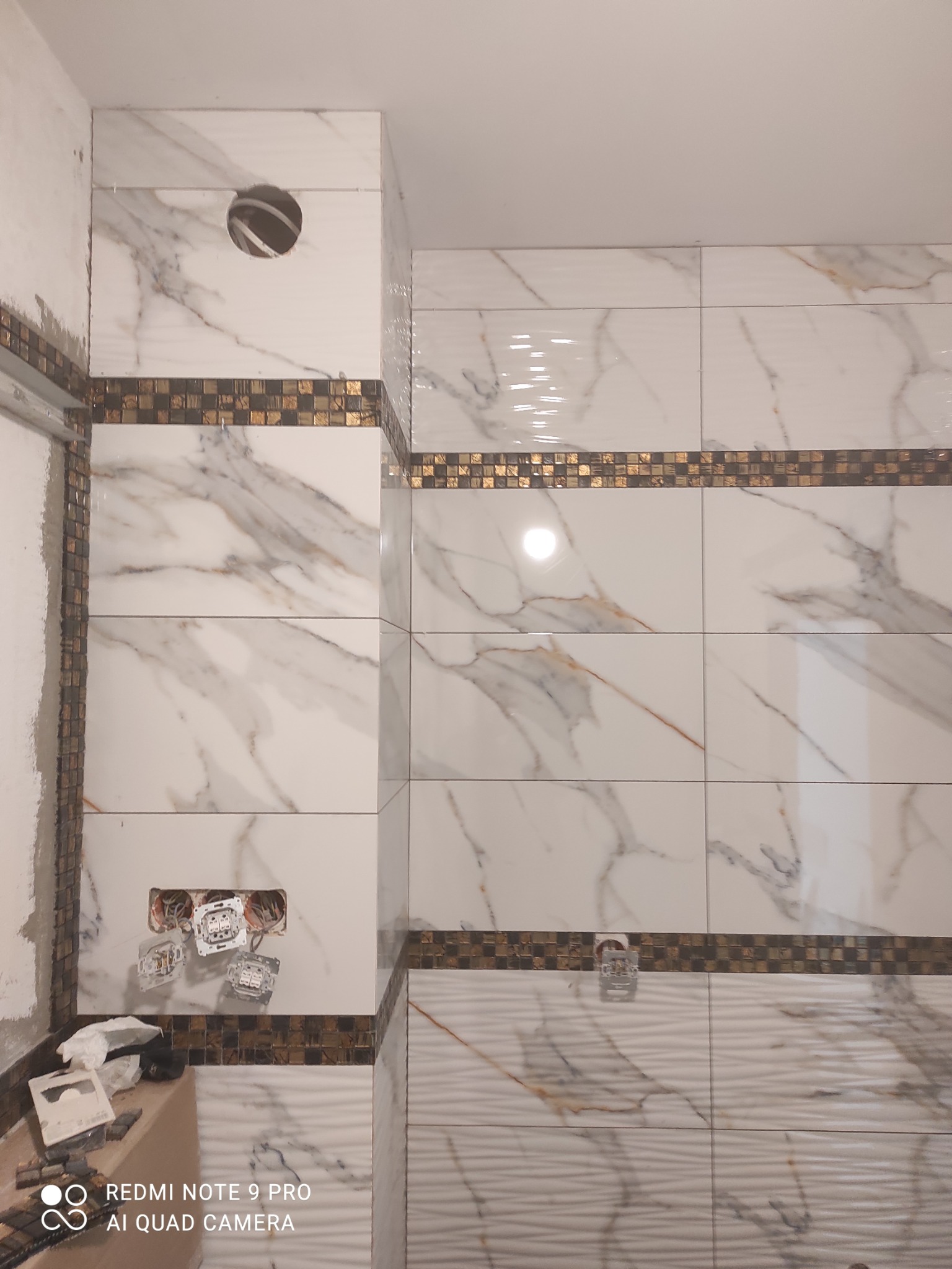 Ściana łazienki w trakcie remontu z białymi płytkami imitującymi marmur, ozdobnymi pasami mozaiki, puszkami elektrycznymi bez osprzętu i narzędziami na podłodze.