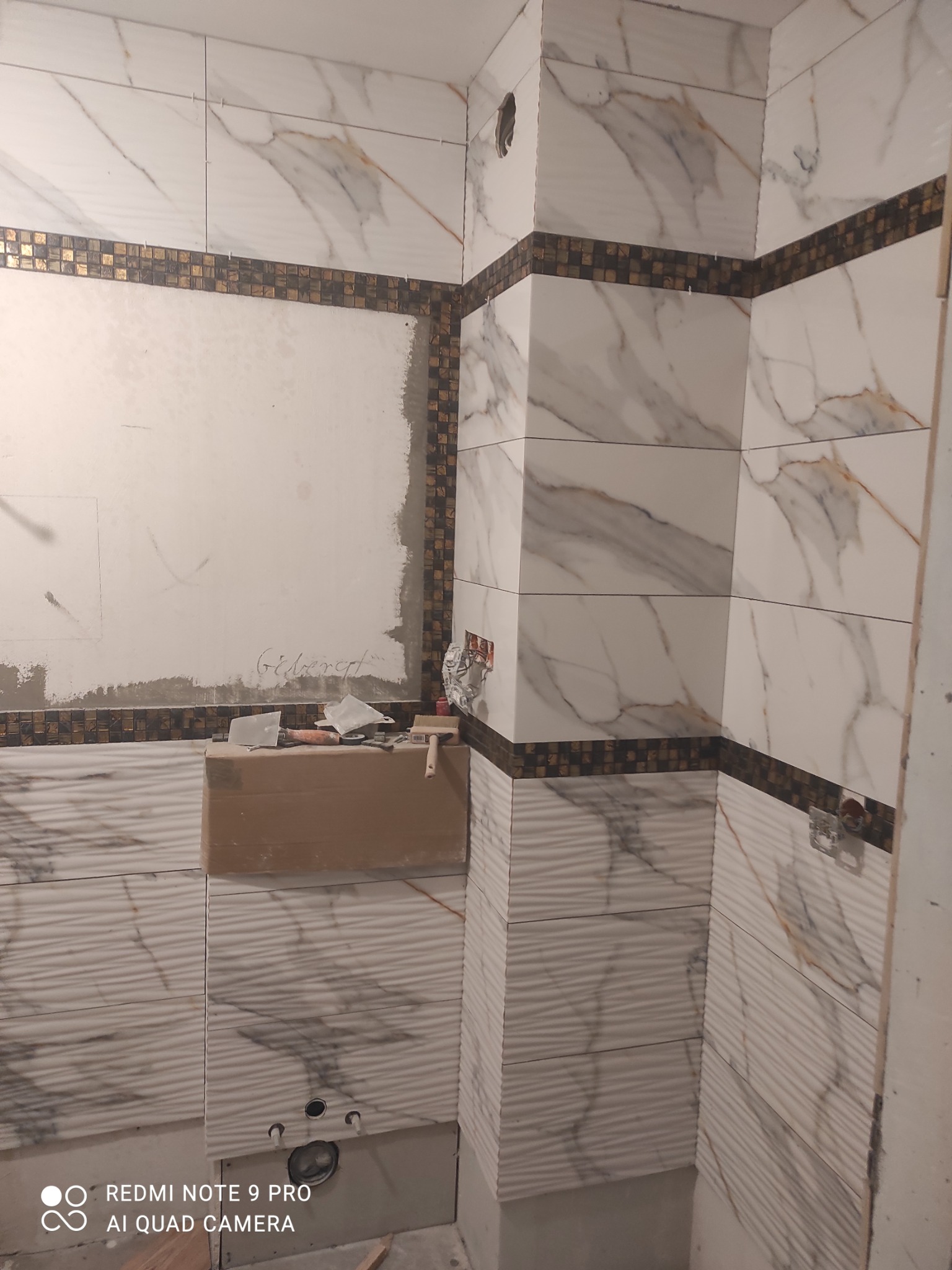 Wnętrze łazienki w trakcie układania płytek imitujących marmur z dekoracyjnym pasem mozaiki, widoczny karton z narzędziami i fragment nieotynkowanej ściany.