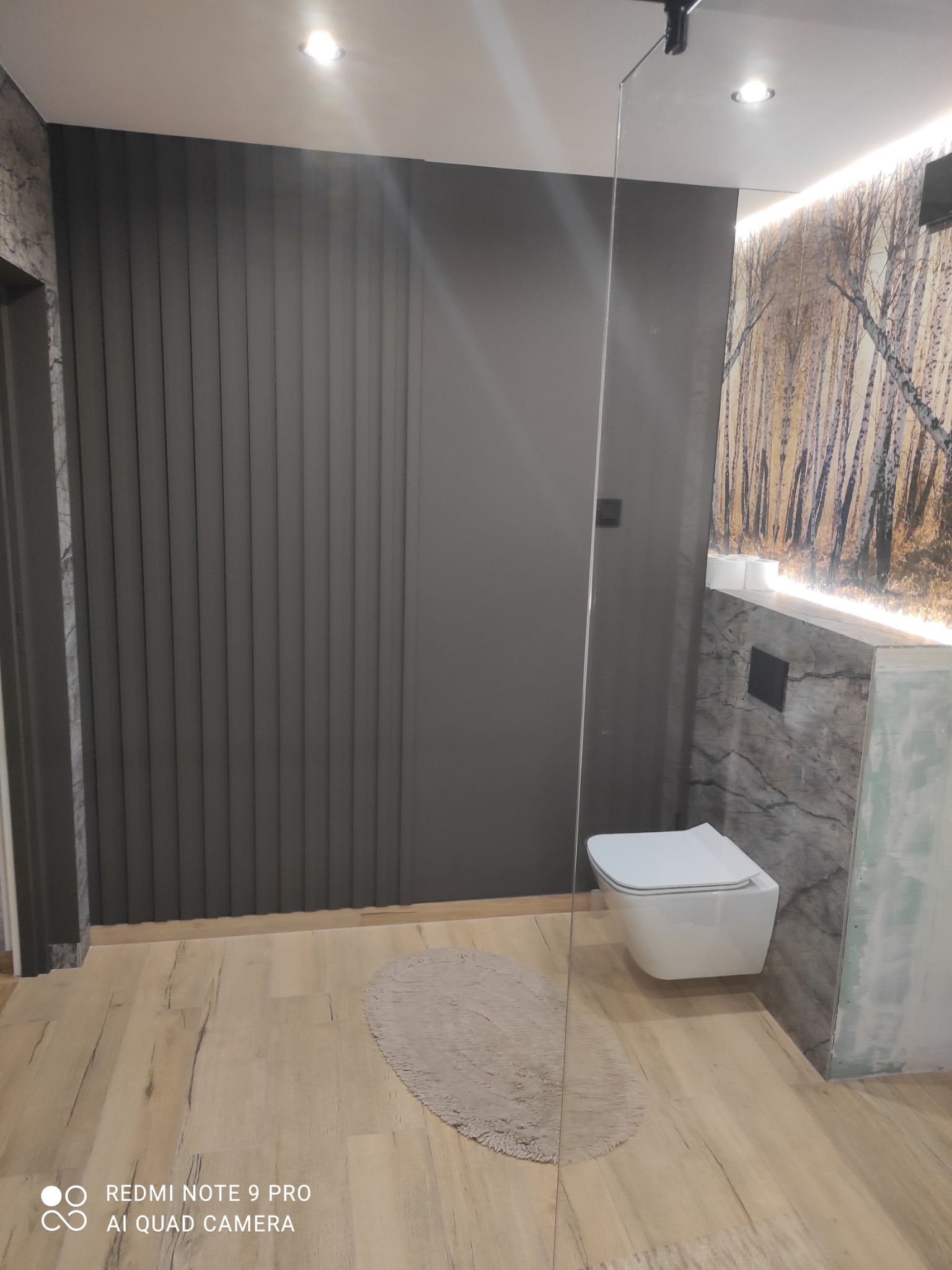 Nowoczesna łazienka z szarą ścianą z pionowymi panelami, podwieszaną toaletą, marmurowym wykończeniem i drewnopodobną podłogą, uzupełniona okrągłym dywanikiem i szklaną ścianką prysznicową.