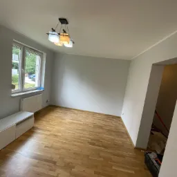 Pusty pokój po remoncie z parkietem, pomalowanymi na szaro ścianami i nowoczesną lampą sufitową; skrzynka z narzędziami widoczna w przejściu do innego pomieszczenia.