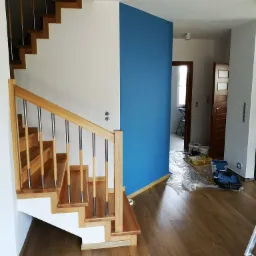 Nowoczesne schody drewniane z metalowymi balustradami w jasnym wnętrzu, widoczne ślady malowania ścian i zabezpieczenia folią.