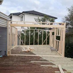 Budowa drewnianej konstrukcji na tarasie, widoczne elementy szkieletu i częściowo wymieniona deska tarasowa, w tle dom jednorodzinny i drzewa.