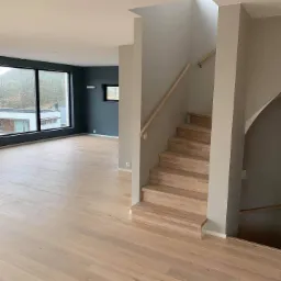 Jasne drewniane schody z poręczą w nowoczesnym wnętrzu domu z dużymi oknami i ciemnymi ścianami.