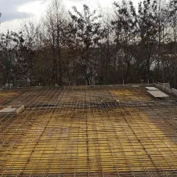 Zbrojenie płyty fundamentowej na żółtym szalunku, widoczne drzewa bez liści w tle.