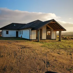 Dom murowany w trakcie budowy z widoczną konstrukcją dachu, ściany z białego bloczka i świeżo położoną dachówką na tle jesiennego krajobrazu.