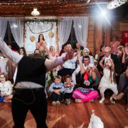 Animator w czarnej kamizelce z uniesionymi rękami przed rozbawioną publicznością podczas występu w drewnianej sali z dekoracjami świetlnymi.