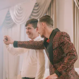 Dwóch mężczyzn w eleganckich strojach, jeden w białej koszuli i muszce, drugi w bordowej marynarce z czarnymi klapami i mikrofonem nagłownym, wykonujących gest dłonią, prawdopodobnie podczas występu.