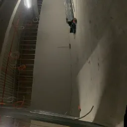 Wnętrze w trakcie prac wykończeniowych: widoczne metalowe profile konstrukcyjne ścian, instalacje elektryczne pod sufitem, podnośnik nożycowy i paleta z płytami na podłodze.