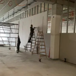 Montaż ściany działowej z płyt gipsowo-kartonowych w surowym stanie, widoczne rusztowanie, drabiny i narzędzia, instalacje pod sufitem