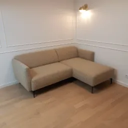 Jasnobrązowa sofa narożna z tapicerką o fakturze baranka, ustawiona w rogu pokoju z białymi ścianami ozdobionymi sztukaterią i drewnianą podłogą, oświetlona złotą lampą ścienną.