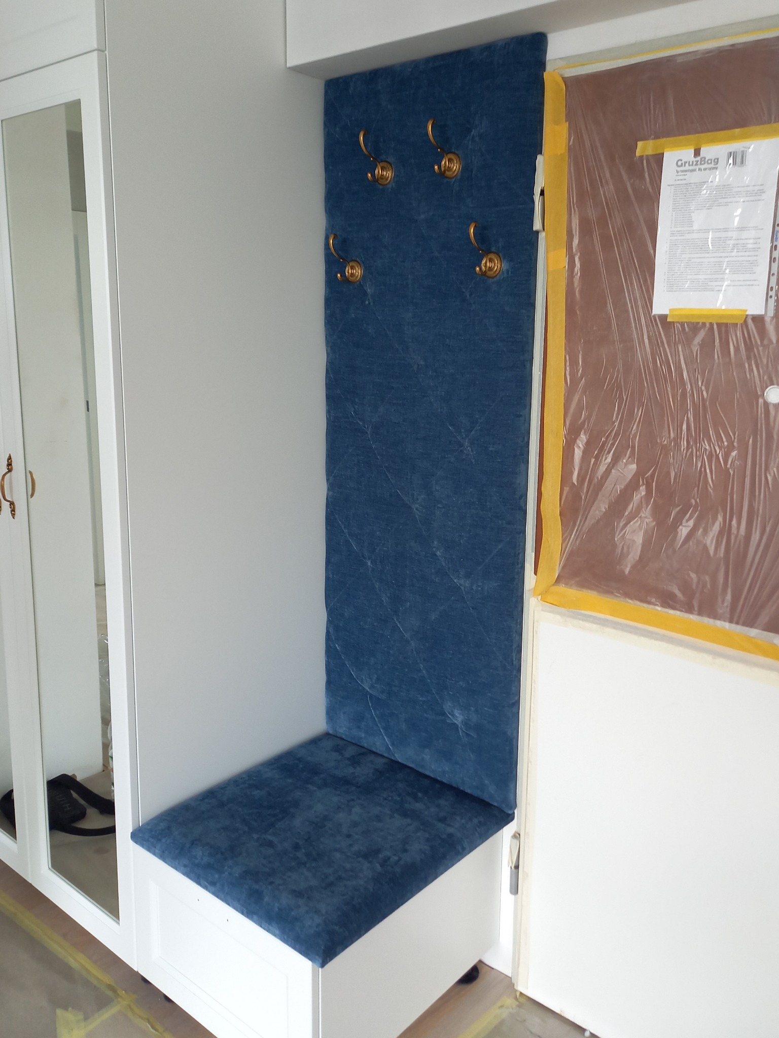 Wnęka w przedpokoju z siedziskiem i panelem ściennym obitym niebieskim, pikowanym welurem, uzupełniona o złote haczyki na ubrania, zamontowana w białej zabudowie meblowej z lustrem, zabezpieczona...