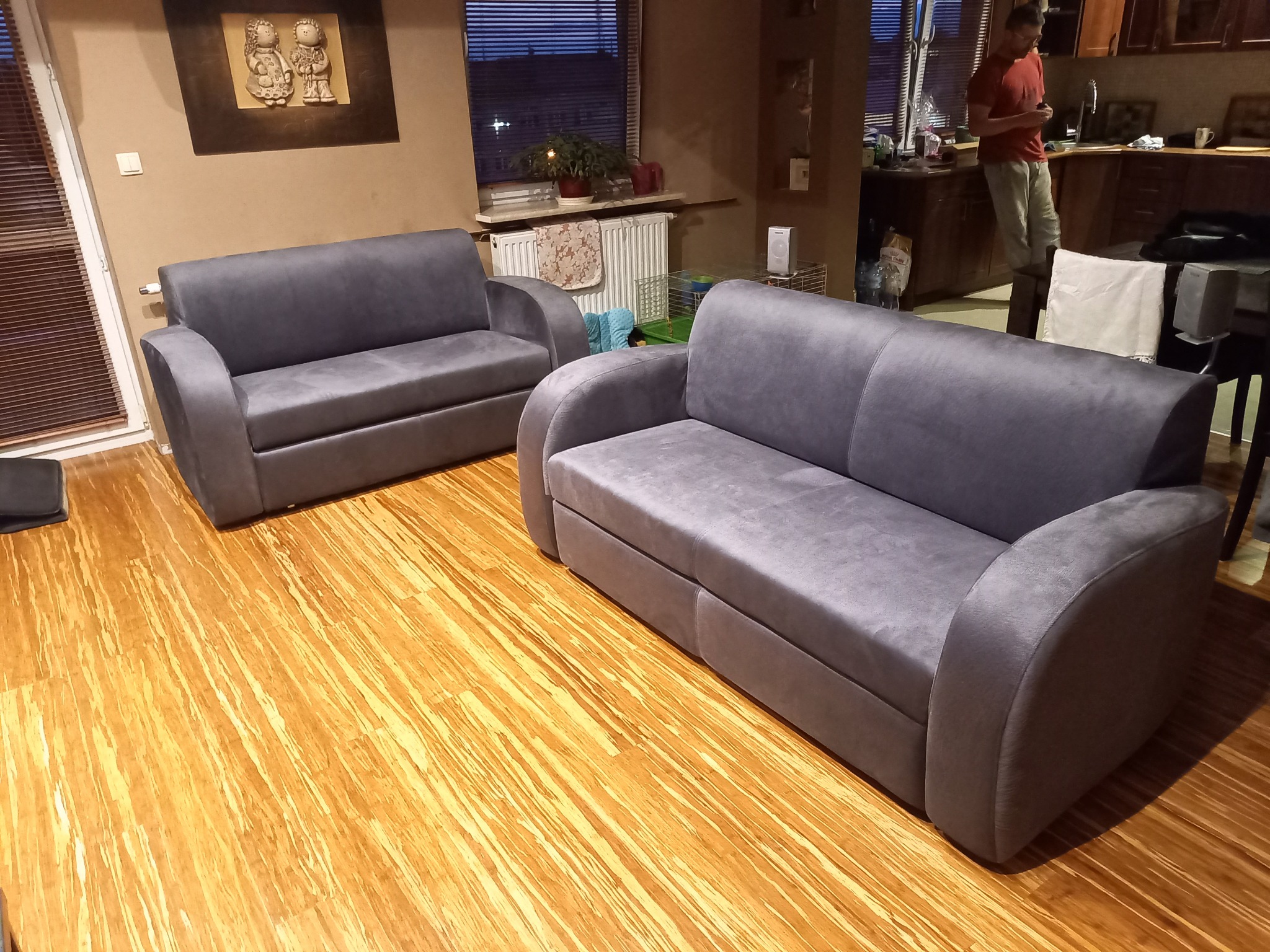 Dwie szare, tapicerowane sofy w salonie na podłodze z paneli bambusowych, z widoczną dekoracją ścienną i osobą w tle.