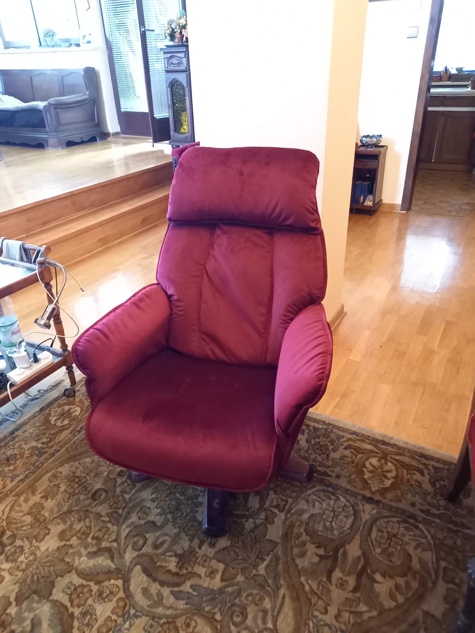 Czerwony, welurowy fotel obrotowy z wysokim oparciem na tle salonu z parkietem i dywanem w orientalne wzory.