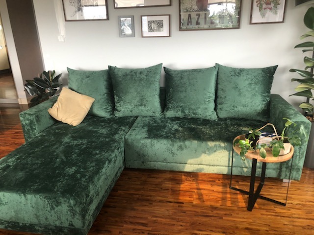 Zielona, welurowa sofa narożna z beżową poduszką, ustawiona na parkiecie, z okrągłym stolikiem z rośliną obok, w jasnym wnętrzu z obrazami na ścianie.