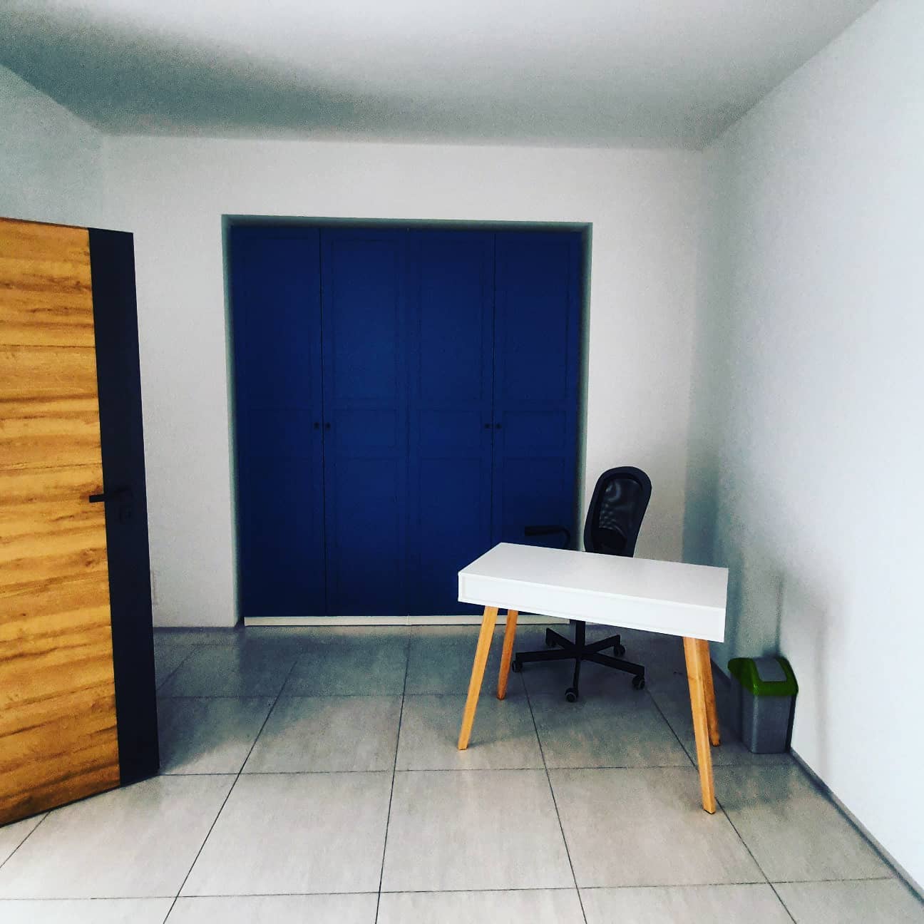 Gabinet logopedy: białe biurko na drewnianych nogach, czarne krzesło biurowe, niebieska szafa wnękowa, drewniane drzwi z czarnymi obramowaniami, szara podłoga z płytek.