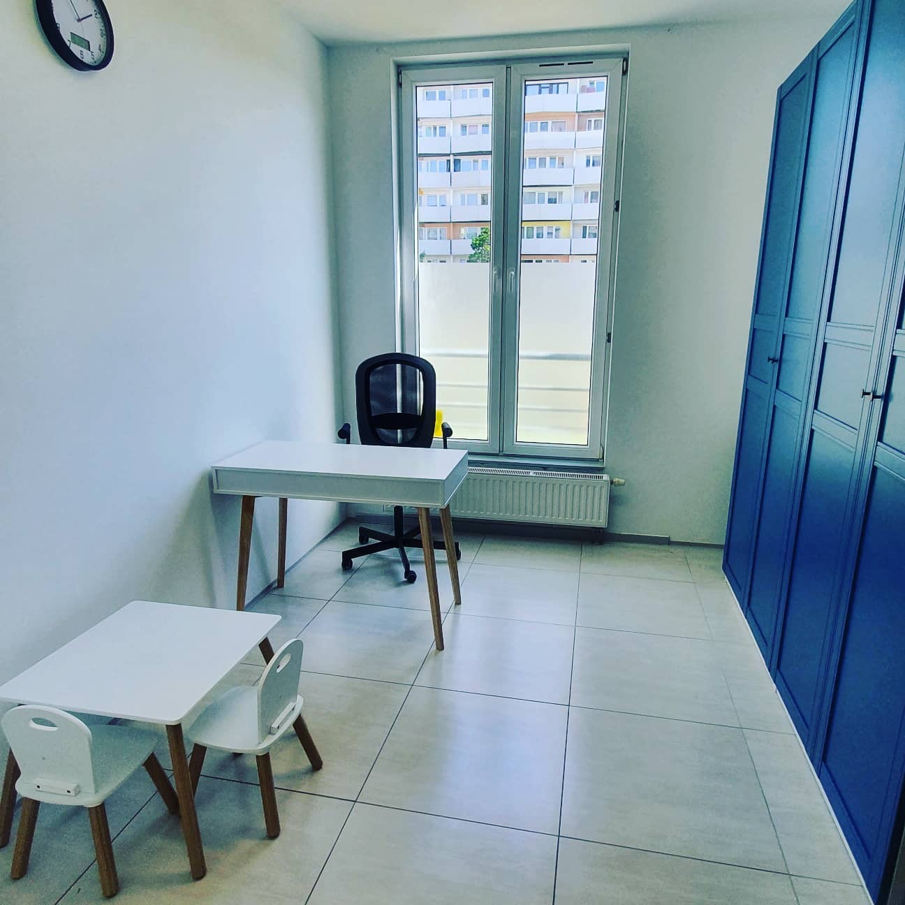 Jasne wnętrze gabinetu z biurkiem dla dorosłych i dzieci, krzesłem biurowym, szafą w kolorze granatowym i oknem z widokiem na blok mieszkalny.