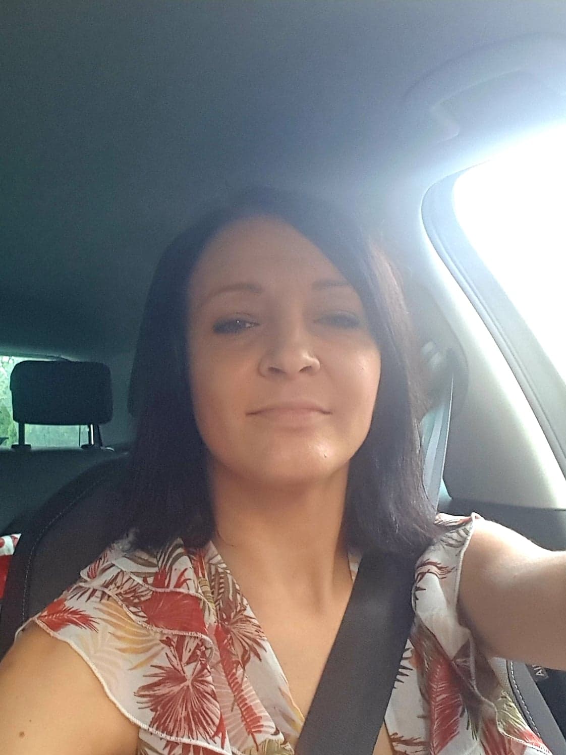 Kobieta z ciemnymi włosami w samochodzie, ubrana w bluzkę w tropikalne wzory, zapięta pasem bezpieczeństwa.