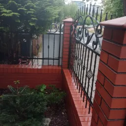 Ceglany murek z kutym, czarnym ogrodzeniem, widoczne ślady wilgoci, zieleń w tle, czerwona szufelka leży na ziemi.