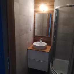 Wykończona łazienka z beżowymi płytkami, drewnianą ścianą za umywalką i kabiną prysznicową z zaokrąglonymi drzwiami.