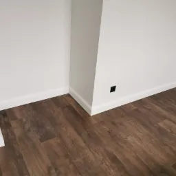 Wnętrze po remoncie: ciemne panele podłogowe z wyraźną strukturą drewna, białe ściany z listwami przypodłogowymi i gniazdko elektryczne. Widoczny narożnik pomieszczenia.