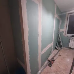 Montaż płyt kartonowo-gipsowych na ścianie, widoczne profile metalowe i szpachlowane łączenia, narzędzia leżące na podłodze z płytek.