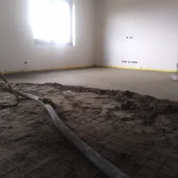 Proces wylewania posadzki: widoczna stalowa siatka z fragmentami świeżej zaprawy, maszyna do zacierania posadzki w tle, oraz wąż doprowadzający mieszankę.