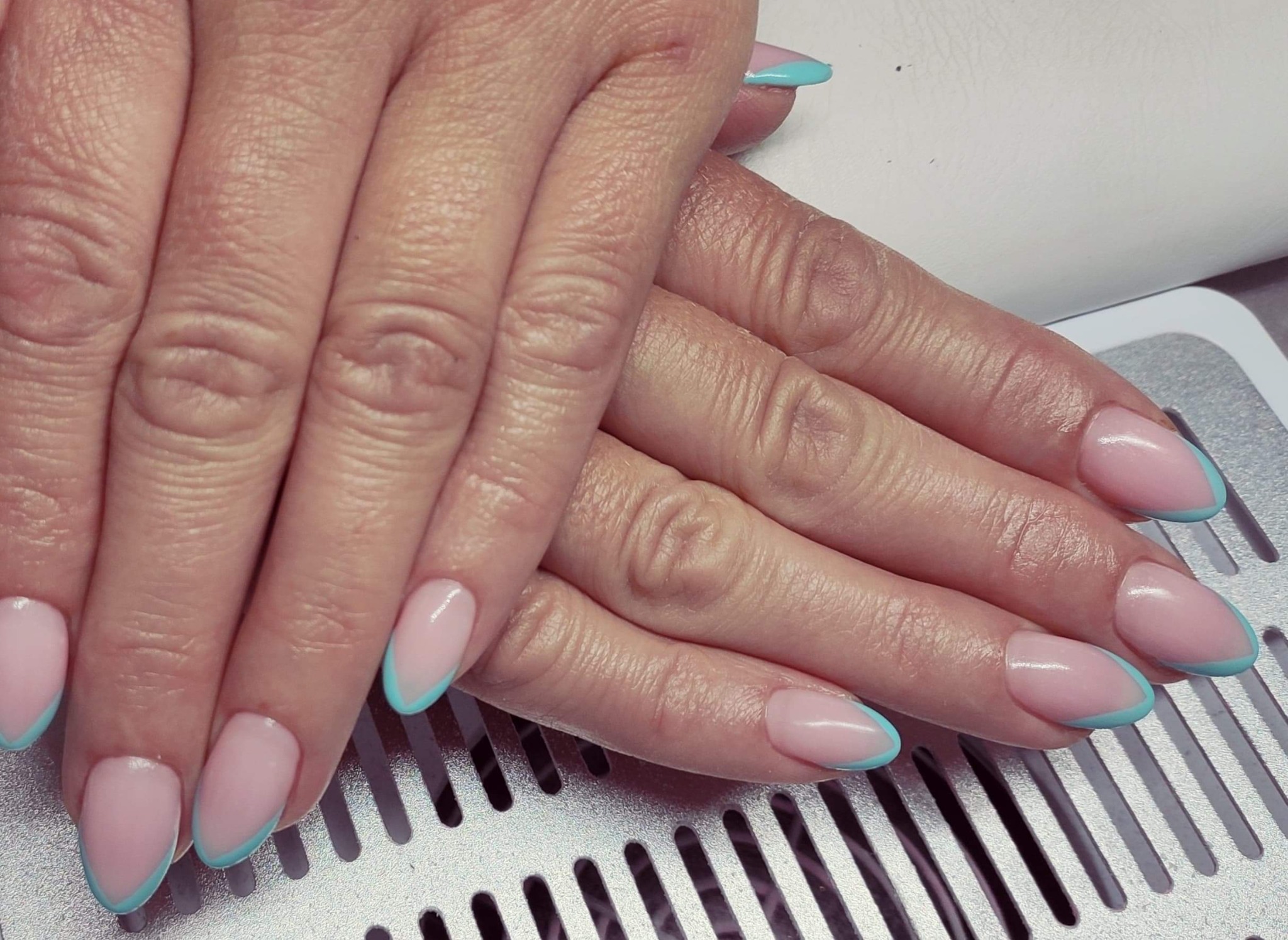 Dłonie z delikatnym manicure hybrydowym: jasnoróżowa baza i turkusowy french na owalnych paznokciach, ułożone na metalowej kratce.