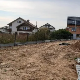 Działka budowlana z widocznym fragmentem ogrodzenia z paneli i betonową podmurówką, w tle domy w różnym stanie wykończenia.