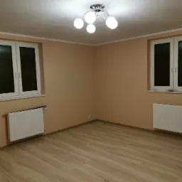 Pomalowane na beżowo puste wnętrze pokoju z drewnianą podłogą, białymi oknami i grzejnikami oraz nowoczesną lampą sufitową.