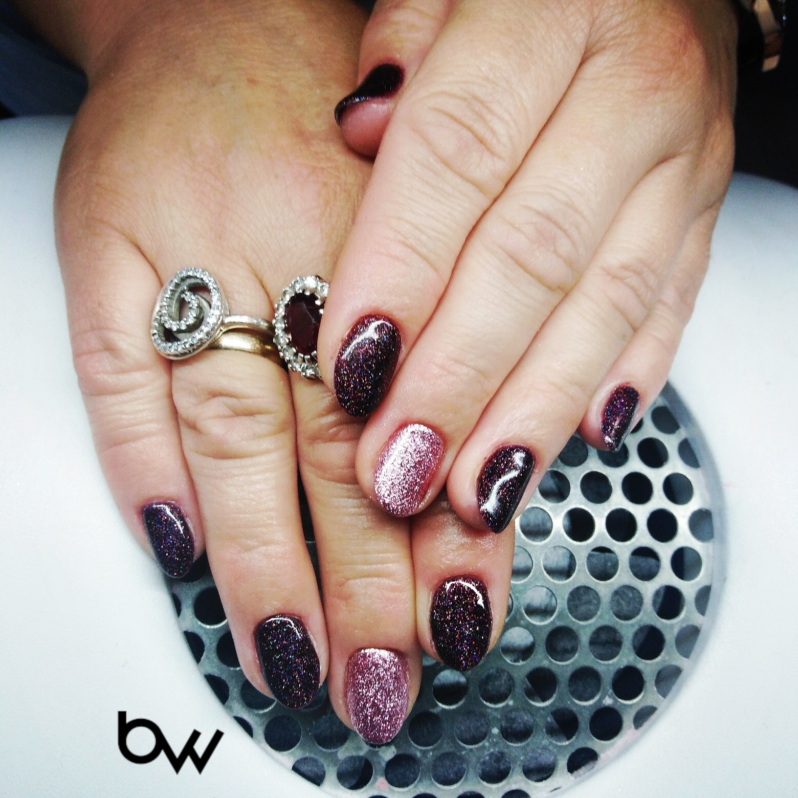 Dłonie z świeżo wykonanym manicure hybrydowym w odcieniach ciemnej śliwki i różowego brokatu, ozdobione pierścionkami, na tle lampy UV.