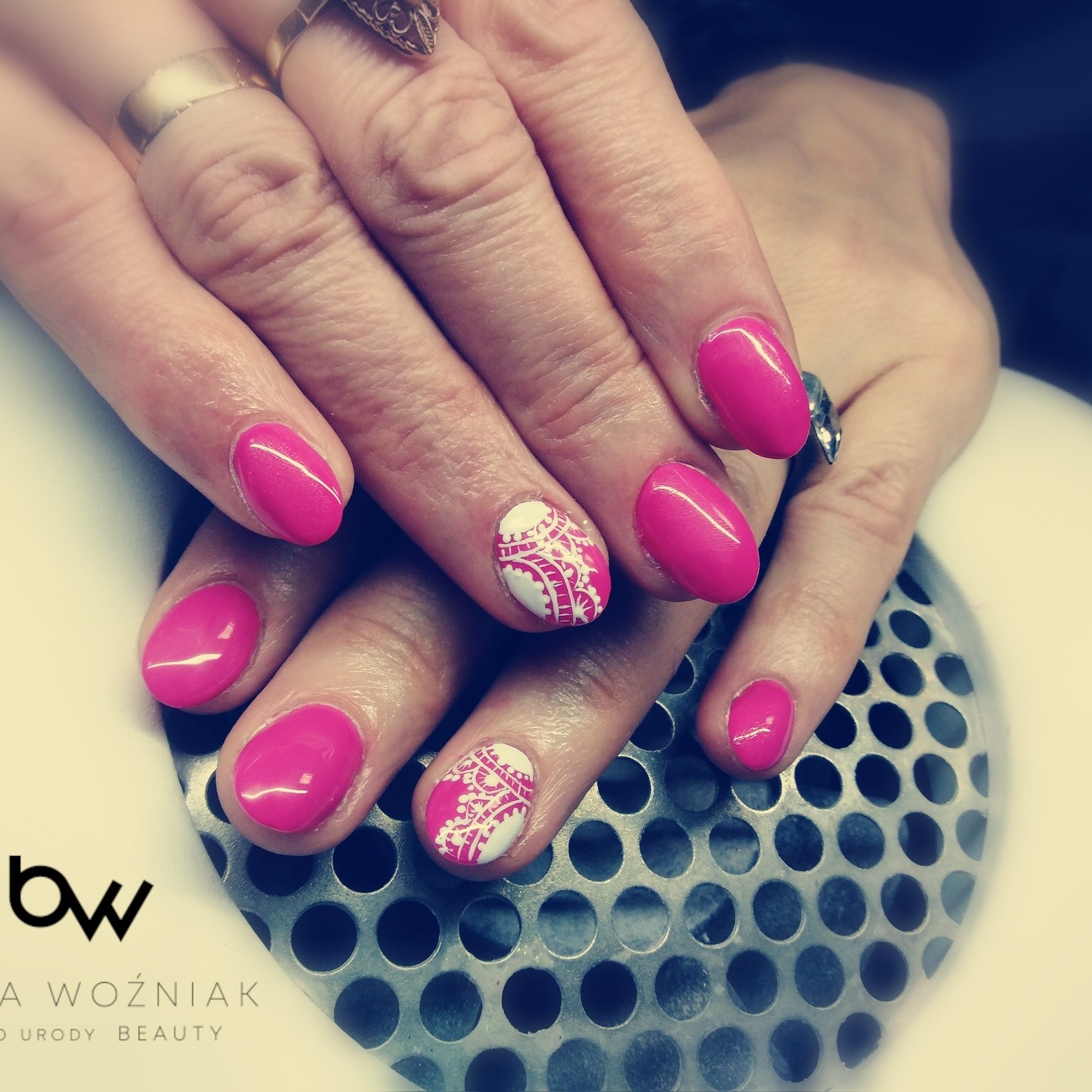 Ręce z paznokciami żelowymi w odcieniach fuksji i białym zdobieniem koronkowym, wykonane w salonie w Łężynach, na tle metalowej kratki.