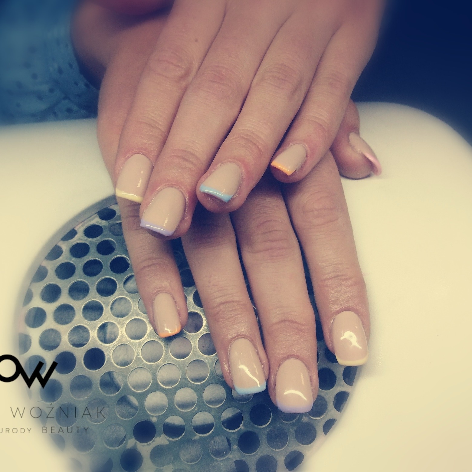 Dłonie z paznokciami żelowymi w odcieniach beżu z pastelowymi końcówkami w kolorach żółtym, niebieskim, pomarańczowym i fioletowym, ułożone na metalowej powierzchni z perforacją.