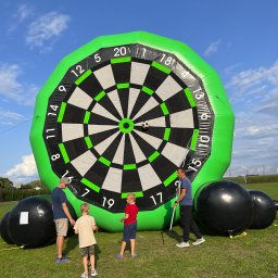 Manufaktura Zabawy - Dmuchana tarcza do gry w rzutki z piłką nożną, otoczona przez dzieci i dorosłych na trawiastym terenie podczas imprezy plenerowej. Widoczne czarne, dmuchane kule.
