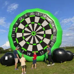 Dmuchana tarcza do gry w rzutki z piłką nożną, otoczona przez dzieci i dorosłych na trawiastym terenie podczas imprezy plenerowej. Widoczne czarne, dmuchane kule.