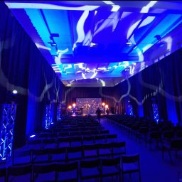 Manufaktura Zabawy - Sala konferencyjna z rzędami krzeseł, oświetleniem scenicznym w kolorze niebieskim i dekoracyjnymi zasłonami. Scena z zespołem muzycznym w tle.