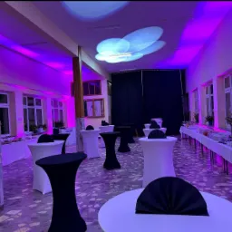 Sala bankietowa z oświetleniem dekoracyjnym, stołami koktajlowymi w czarnych i białych pokrowcach oraz bufetem z napojami i przekąskami, gotowa na przyjęcie.