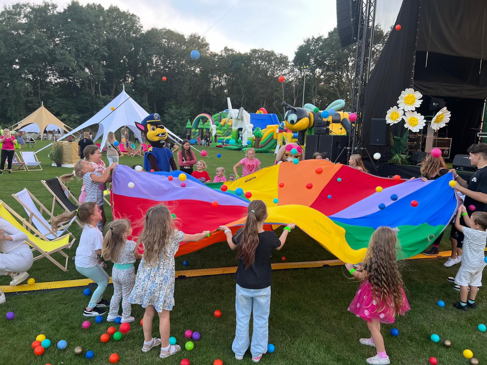 Kolorowa zabawa z animatorem w stroju, dmuchańce w tle. Dzieci trzymają tęczową płachtę z piłeczkami na trawie, scena i namioty w tle.