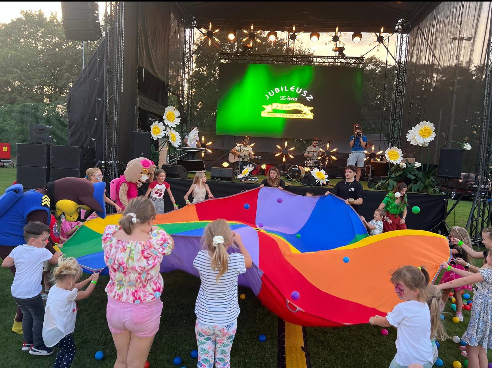 Plenerowa impreza z okazji jubileuszu: scena z zespołem, animatorzy w kostiumach i dzieci bawiące się kolorową chustą animacyjną na trawie.