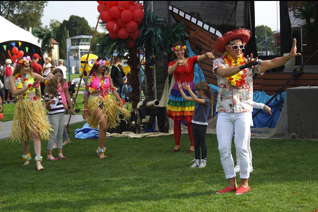 Egzotyczna impreza plenerowa: osoby w strojach hawajskich tańczą na trawie, w tle dekoracje tematyczne, w tym armata i balony. Mężczyzna w kapeluszu z mikrofonem prowadzi zabawę.