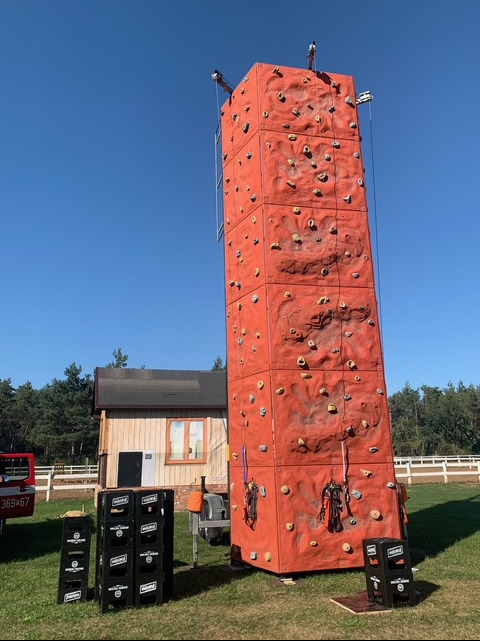Mobilna ścianka wspinaczkowa w kolorze pomarańczowym na przyczepie, ustawiona na trawie, z widocznym sprzętem wspinaczkowym i stertą skrzynek w tle.