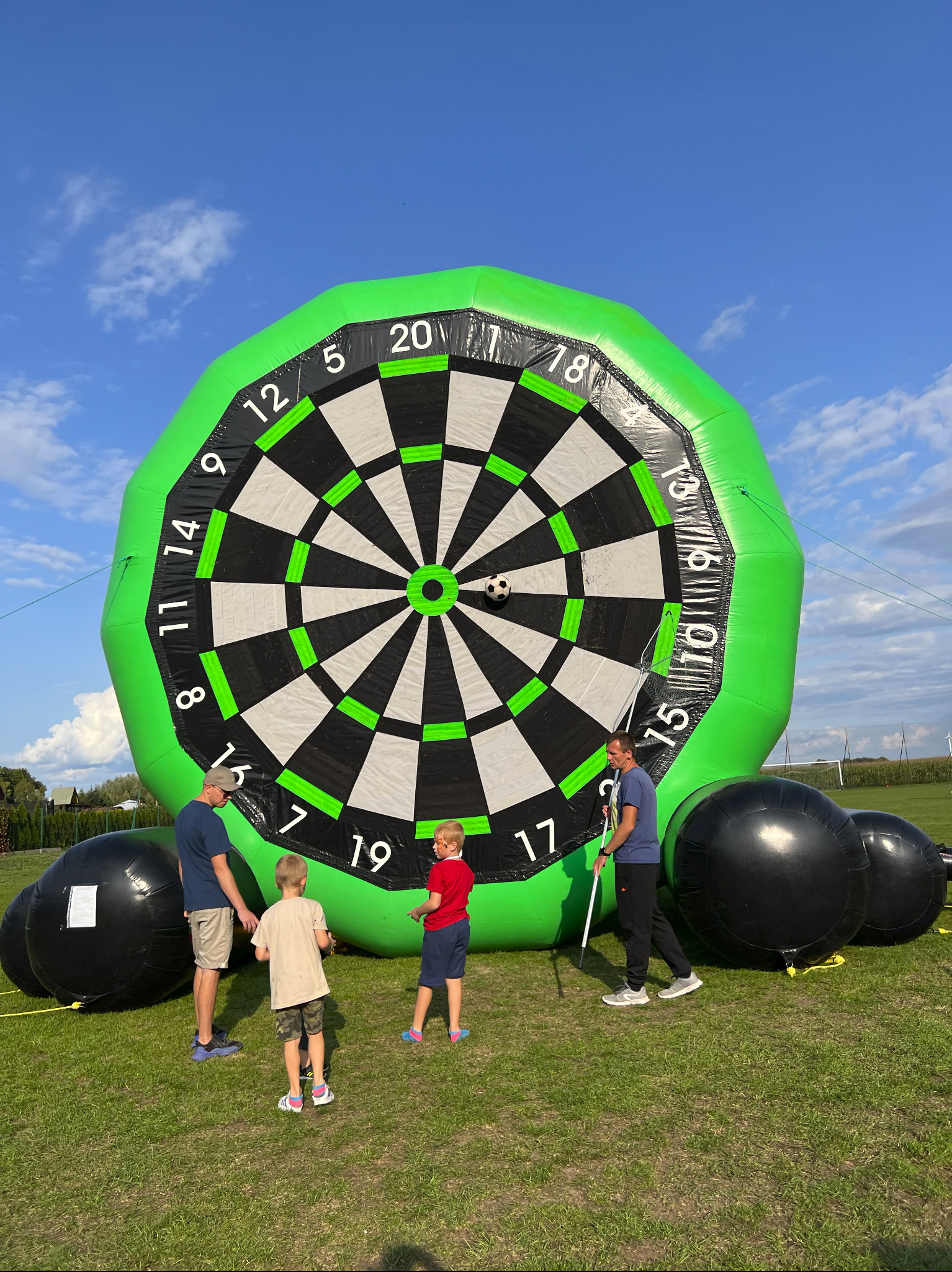 Dmuchana tarcza do gry w rzutki z piłką nożną, otoczona przez dzieci i dorosłych na trawiastym terenie podczas imprezy plenerowej. Widoczne czarne, dmuchane kule.