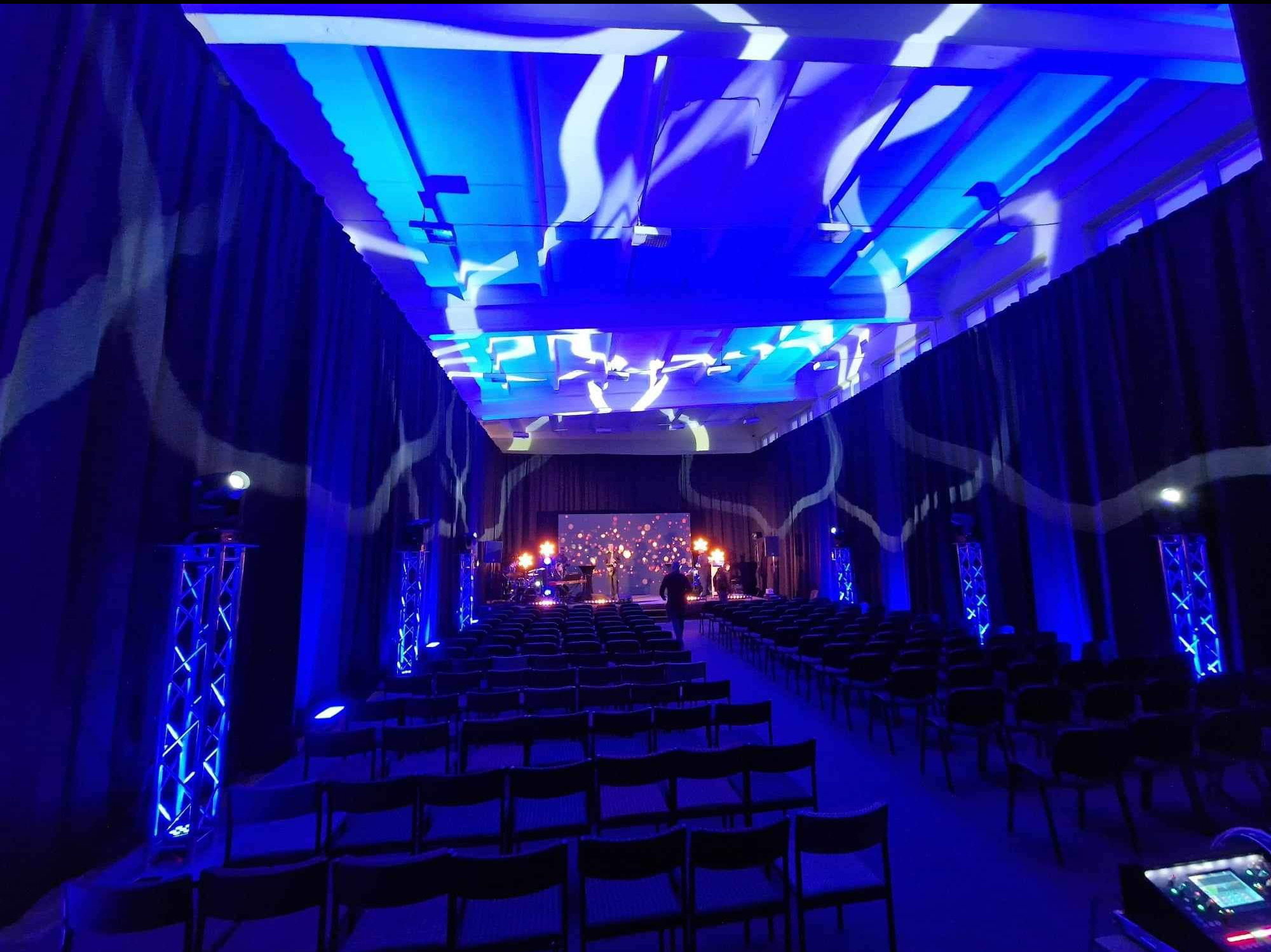 Sala konferencyjna z rzędami krzeseł, oświetleniem scenicznym w kolorze niebieskim i dekoracyjnymi zasłonami. Scena z zespołem muzycznym w tle.