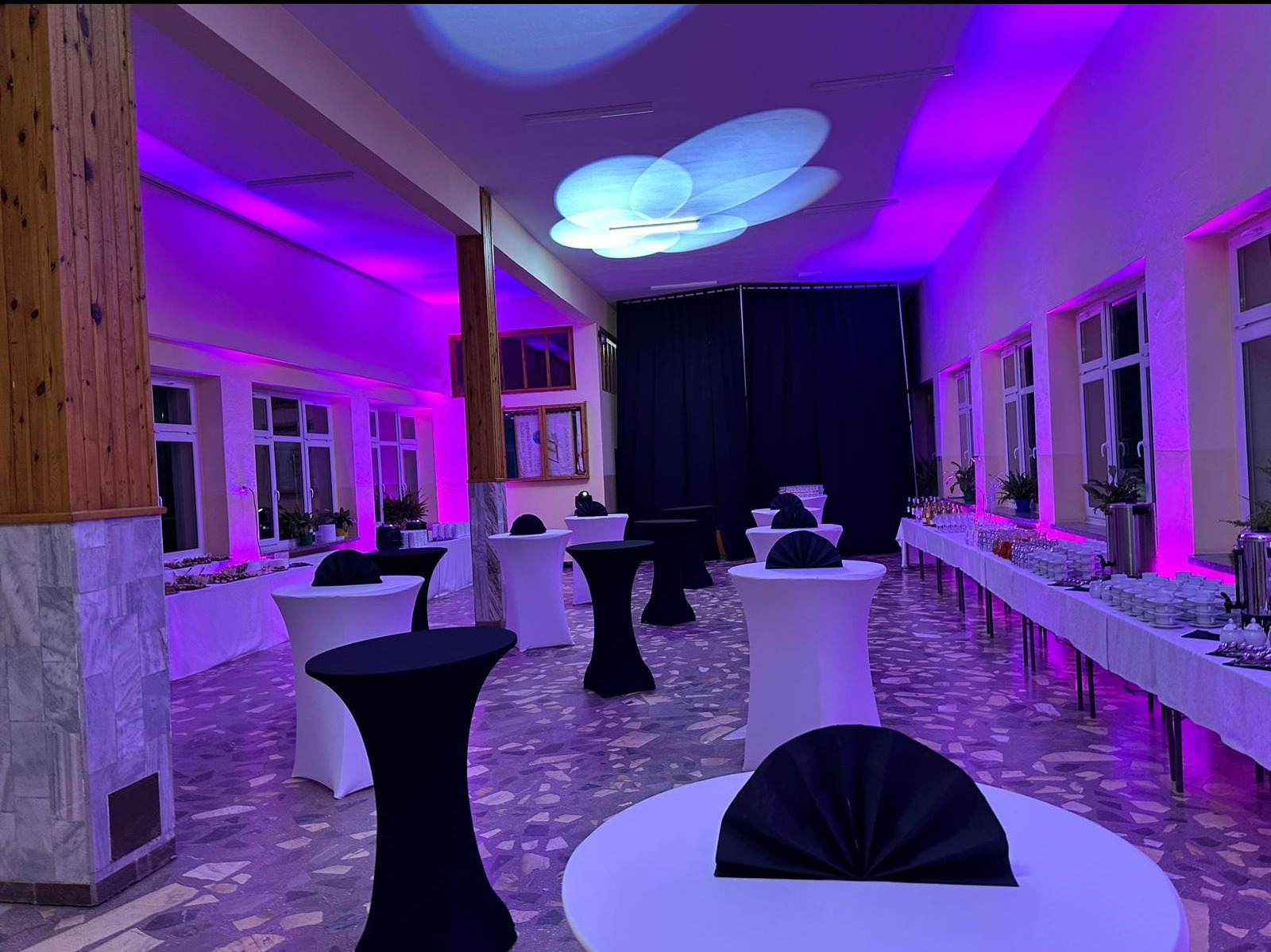 Sala bankietowa z oświetleniem dekoracyjnym, stołami koktajlowymi w czarnych i białych pokrowcach oraz bufetem z napojami i przekąskami, gotowa na przyjęcie.